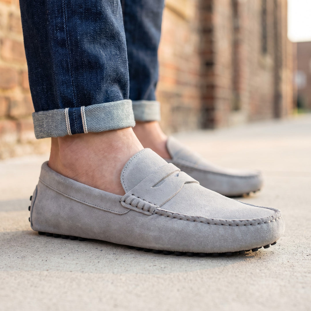 mocassin en daim gris clair porté avec un jean brut à revers sur un trottoir devant un mur de brique